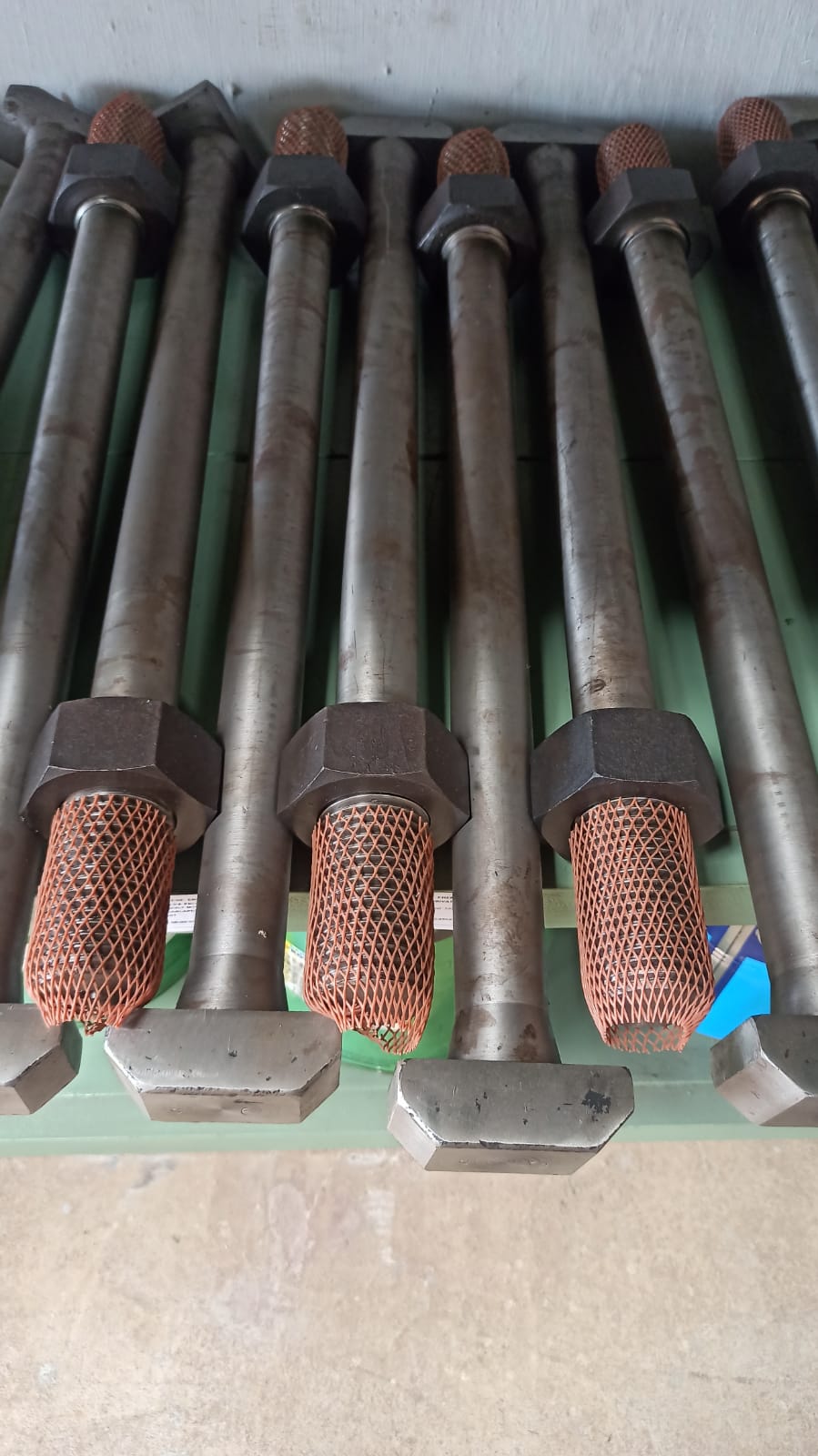 Row of long T-head bolts with threaded ends protected by plastic mesh and secured with heavy hex nuts